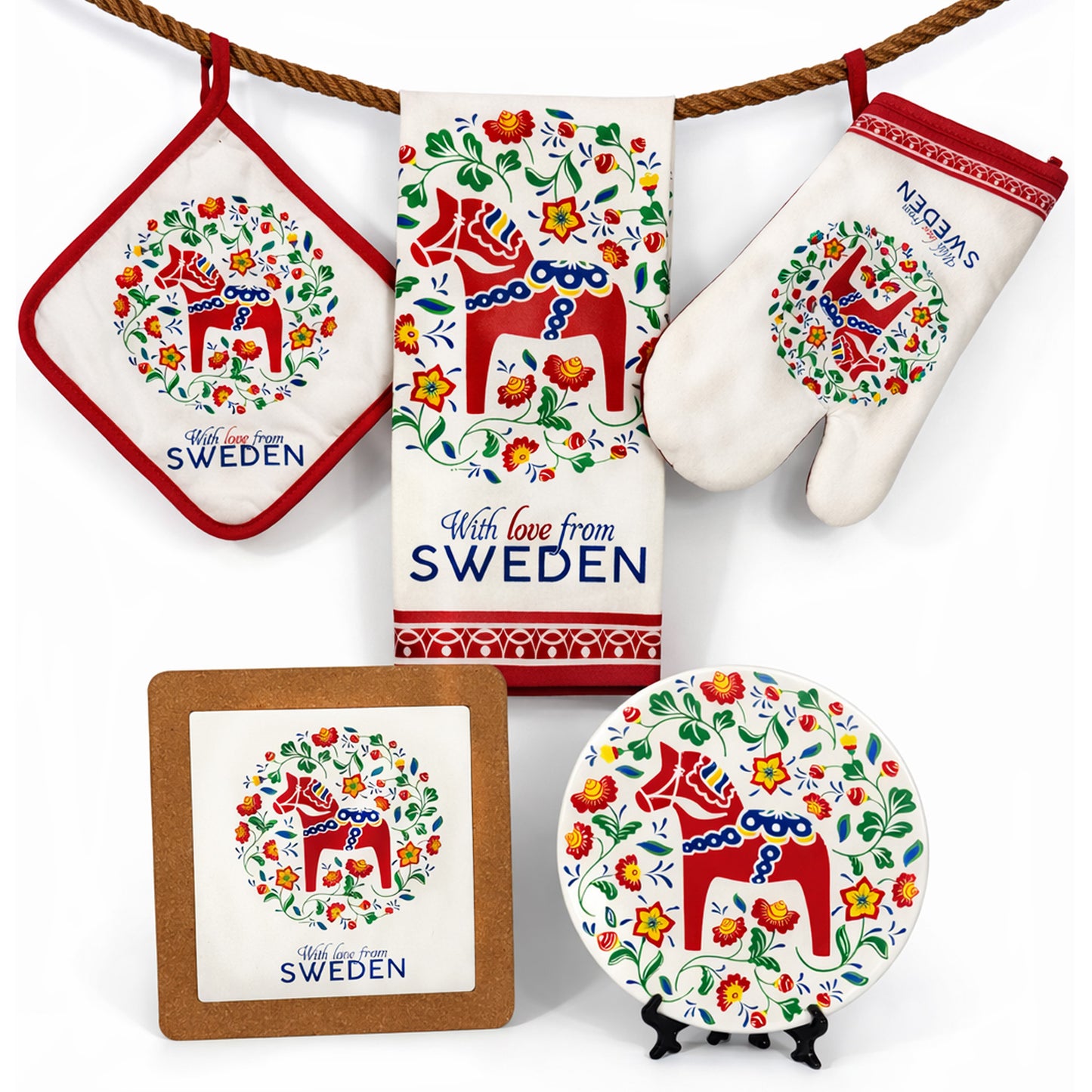 Ett matchande set med svensk-inspirerade kökstextilier och dekorationer som hänger på ett rep. Det består av en grytlapp, en kökshandduk och en grytvante – alla med en röd dalahäst omgiven av färgglada blommönster och texten ”With love from Sweden”. Nedanför syns även ett matchande grytunderlägg och en dekorativ tallrik med samma dalahästmotiv.