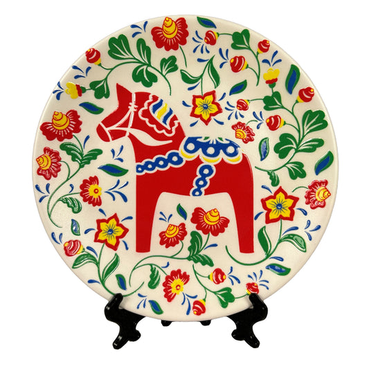Rund souvenirtallrik prydd av en färgglad dalahäst som är omgiven av snirklande blommor. Tallriken kommer med både ställ och väggfäste.
