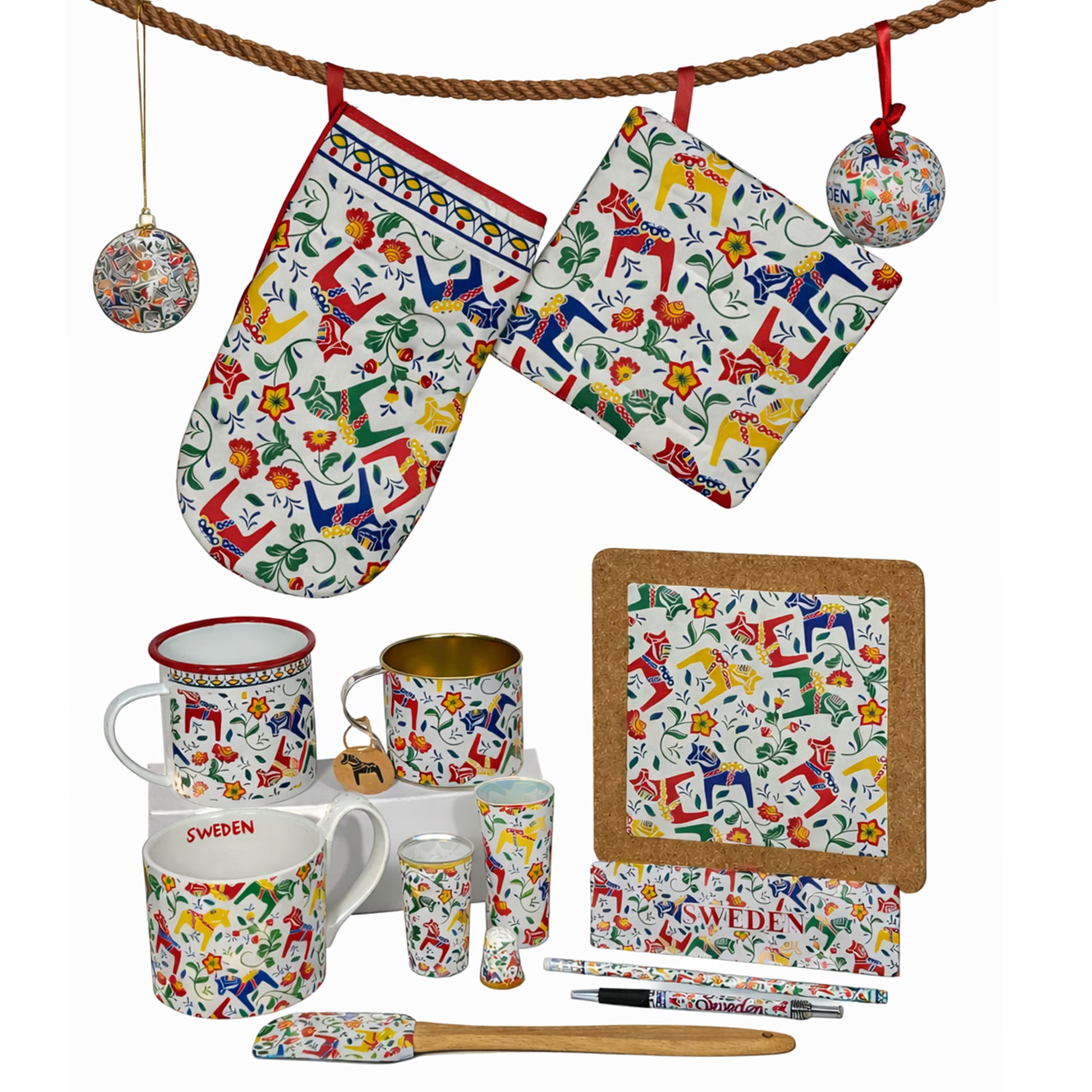 En dalahäst inspirerad souvenirserie med grytlapp, grytunderlägg och juldekorationer hängandes på ett brunt rep. Nedanför står muggar, shotglas, fingerborg, magnet, pennor och en slickepott – alla med samma färgstarka motiv av dalahästar omgivna av snirklande blommor.