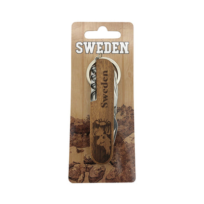 Ihopfälld fällkniv i trä med älgmotiv och texten "Sweden", fäst på en hängetikett med skogsmotiv och texten "Sweden". 