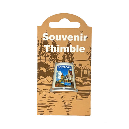 Vit fingerborg med motiv från staden Göteborg . Visas här i sin förpackning = en ljusbrun hängetikett med mörkbrunt skogsmotiv och texten Souvenir Thimble i vitt. 