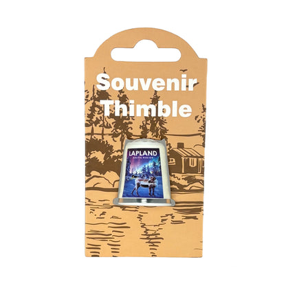Fingerborg med vintermotiv från Lappland – ståtlig ren i snölandskap med norrsken i bakgrunden. Visas här i sin förpackning = ljusbrun hängetikett med mörkbrunt skogsmotiv + texten Souvenir Thimble i vitt. 