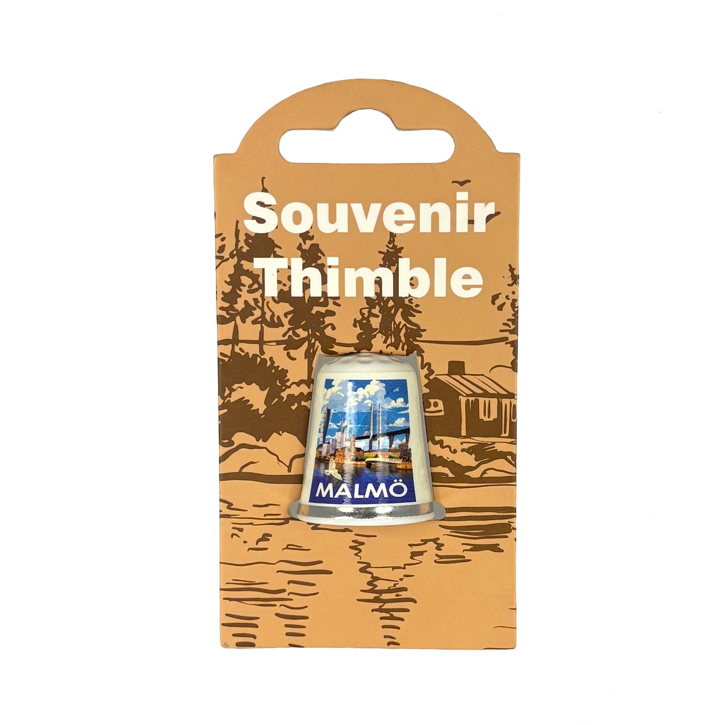 Vit fingerborg med motiv från Malmö. Visas här i sin förpackning = ljusbrun hängetikett med mörkbrunt skogsmotiv och texten "Souvenir Thimble" i vitt. 