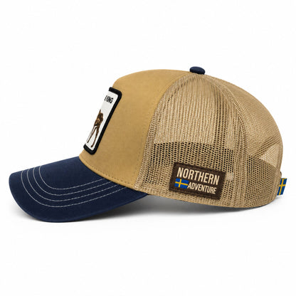 Ljusbrun och blå truckerkeps sedd från sidan. Här syns ett märke med texten “Northern Adventure” och  ensvensk flagga – en snygg keps med älgmotiv och justerbar nätbakdel.