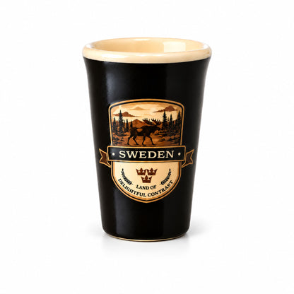 Svart shotglas med crèmefärgad insida och ett 2D-motiv av en älg i en skog, samt texten "Sweden – Land of Delightful Contrast".