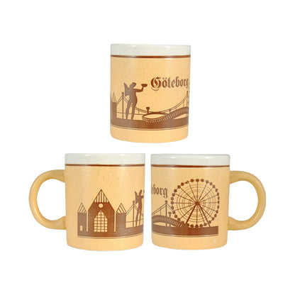 Keramikmugg med Göteborgsmotiv sedd från tre vinklar – med graverade motiv av Poseidon, Ullevi, Älvsborgsbron, pariserhjulet på Liseberg, Feskekörka och texten "Göteborg". Vit insida och stilren souvenirkänsla.