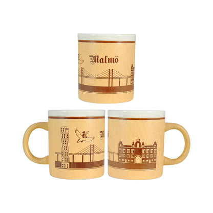 Keramikmugg med Malmömotiv sedd från tre vinklar – med graverade motiv av Öresundsbron, Turning Torso, Centralstationen, texten "Malmö" och en liten Nils Holgersson på gås. En gedigen souvenir med vit insida.