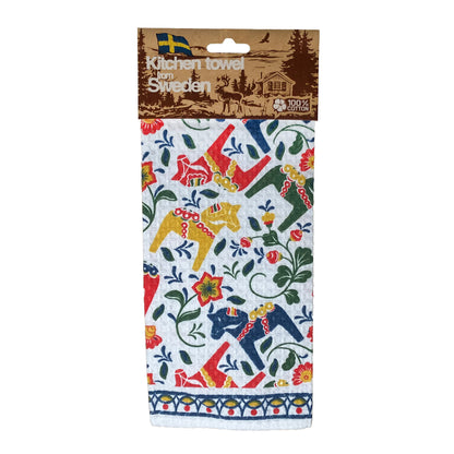 Färgglad kökshandduk med dalahästar omgivna av fina blommor. Här visas handduken med ljusbrun hängetikett med mörkbrunt skogsmotiv med en ren och liten stuga, texten "Kitchen Towel from Sweden" och "100 % cotton".