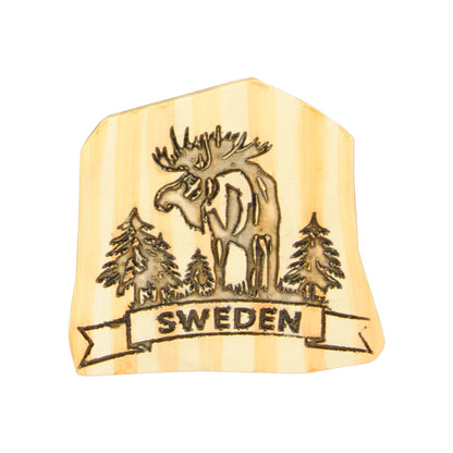 Souvenirmagnet med graverat motiv av stående älgtjur och skog samt texten "Sweden". Ytan har ett ljust träliknande utseende som ger ett naturligt och exklusivt intryck.
