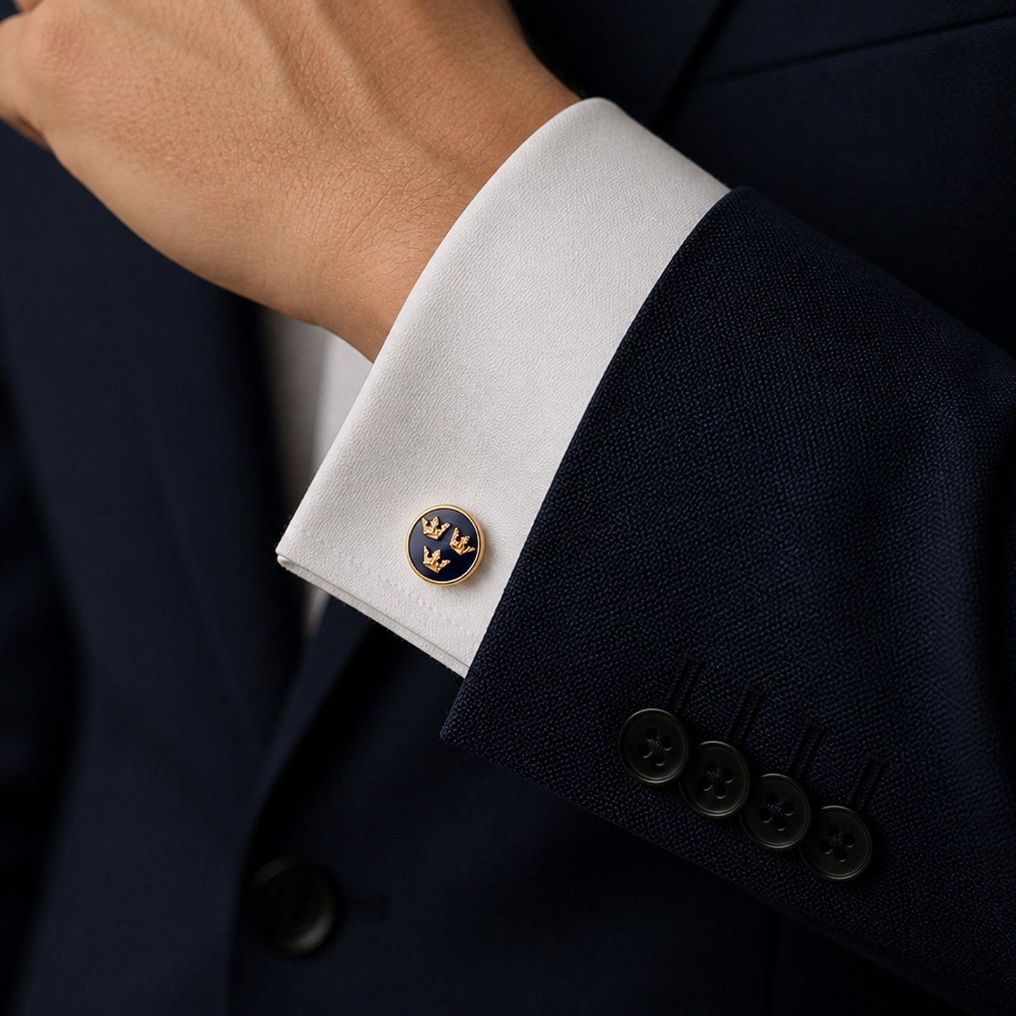 Guldfärgade manschettknapp med blå emalj och Tre Kronor-detalj, elegant placerade på vit skjortärm tillsammans med mörk kostymärm – en klassisk och stilren accessoar för formella tillfällen.