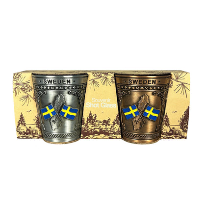 2-pack metall shotglas i silver och brons. Kanten upptill pryds av texten SWEDEN medan glasens framsida pryds av tre klassiska svenska motiv: Sverige kartan med två korsande flaggor, ett historiskt vikingaskepp och ett ståtligt älghuvud. Visas här i sin förpackning med texten "Souvenir Shot Glass".