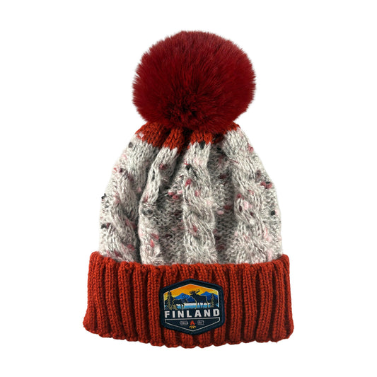 Stickad röd och grå mössa med röd tofs och ett märke med texten Finland på mudden - sedd framifrån. 