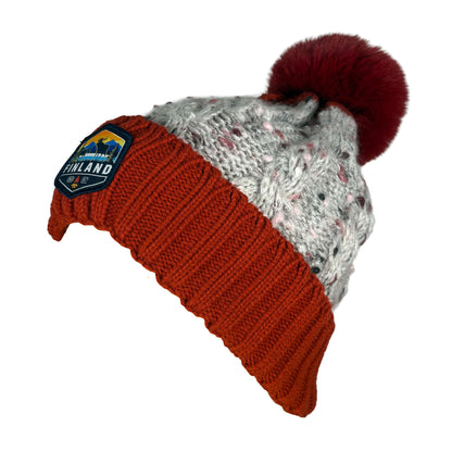 Stickad röd och grå mössa med röd tofs och ett märke med texten Finland på mudden - sedd från sidan. 