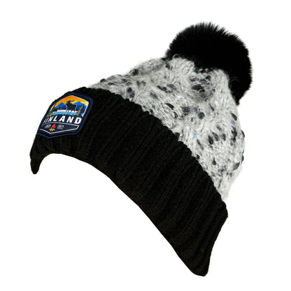 Stickad svart mössa med svart tofs och märke med texten Finland på mudden- sedd från sidan. 