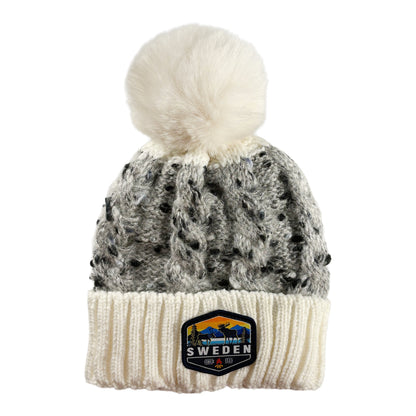 Stickad mössa i vitt och grått med fluffig vit tofs, sedd framifrån. Vävt märke med älgmotiv och texten "Sweden" synligt på mudden.