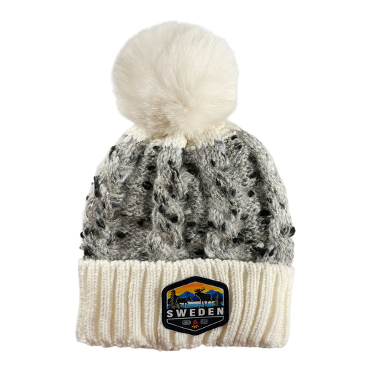 Stickad mössa i vitt och grått med fluffig vit tofs, sedd framifrån. Vävt märke med älgmotiv och texten "Sweden" synligt på mudden.