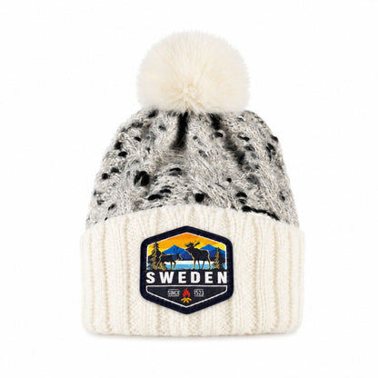 Stickad mössa i vitt och grått med fluffig vit tofs, sedd framifrån. Vävt märke med älgmotiv och texten "Sweden" synligt på mudden.