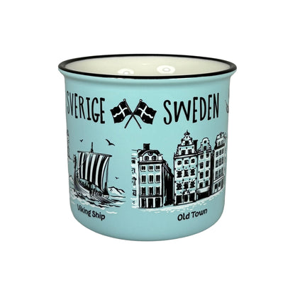Ljusblå mugg utan synligt handtag med svartvita illustrationer av två korsade svenska flaggor, ett vikingaskepp, Gamla Stan och texten 'Sverige Sweden'.