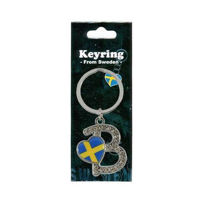 Metallnyckelring med bokstaven B. Bokstaven är dekorerad med glitterstenar. På bokstaven finns även ett hjärta med en svensk flagga. Visas här i sin förpackning. Svart förpackning med texten "Keyring from Sweden" i vitt. 