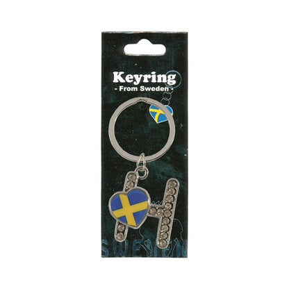 Metallnyckelring med bokstaven H. Bokstaven är dekorerad med glitterstenar. På bokstaven finns även ett hjärta med en svensk flagga. Visas här i sin förpackning. Svart förpackning med  texten "Keyring från Sweden" i vitt.