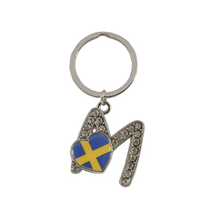 Metallnyckelring med bokstaven M. Bokstaven är dekorerad med glitterstenar. På bokstaven finns även ett hjärta med en svensk flagga.