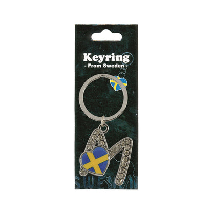 Metallnyckelring med bokstaven M. Bokstaven är dekorerad med glitterstenar. På bokstaven finns även ett hjärta med en svensk flagga. Visas här i sin förpackning. Svart förpackning med  texten "Keyring från Sweden" i vitt.