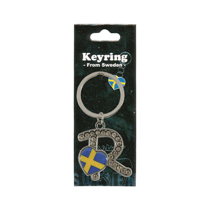 Metallnyckelring med bokstaven R. Bokstaven är dekorerad med glitterstenar. På bokstaven finns även ett hjärta med en svensk flagga. Visas här i sin förpackning. Svart förpackning med  texten "Keyring från Sweden" i vitt.