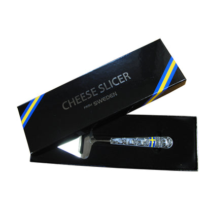 Osthyvel i metall med utskurna svenska motiv, i svart presentask med glågult band och texten 'Cheese slicer from Sweden'.
