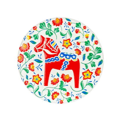 Rund magnet med röd dalahäst omgiven av blomster och gröna blad, en färgglad svensk souvenir.