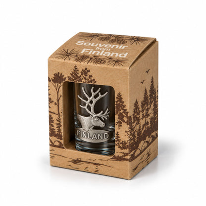 Elegant shotglas prytt med ett metallmärke i form av en ren och texten "Finland", visas i sin förpackning - snett framifrån.
