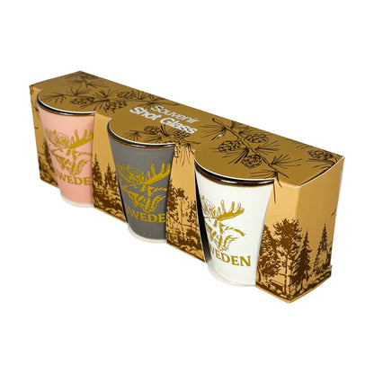 Tre shotglas ett rosa, ett vitt och ett grått. Alla med guldfärgat älghuvud och texten "Sweden". Visas här snett uppifrån i sin förpackning. 