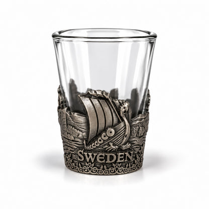Shotsglas i glas med ett omslag i tennfärgad metall med vikingamotiv. Här syns ett vikingaskepp och texten 