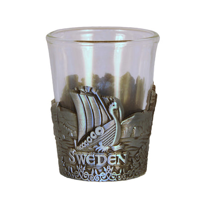 Shotsglas i glas med ett omslag i tennfärgad metall med vikingamotiv. Här syns ett vikingaskepp och texten Sweden. 