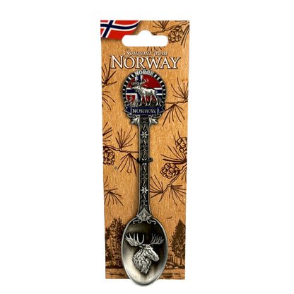 Silverfärgad souvenirsked med ett älghuvud i skedens kupa. Skaftet är dekorerat med ett fint mönster, en älg, norska flaggan samt texten "Norge" och "Norway". Visas här på ljusbrun hängetikett prydd med ett mörkbrunt skogsmotiv, norska flagga och texten "Souvenir from Norway".