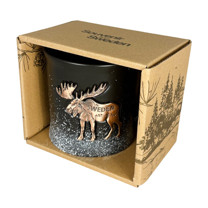 Svart mugg med bronsfärgad älg i en brun pappförpackning med mörkbrunt skogsmotiv och texten "Souvenir from Sweden" på toppen