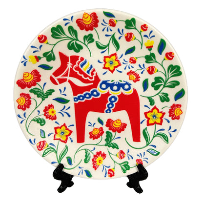 Rund souvenirtallrik prydd av en färgglad dalahäst som är omgiven av snirklande blommor. Visas här med ställ.