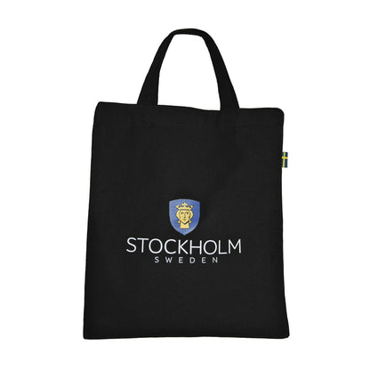 Baksida av svart tygkasse dekoerad med texten "STOCHOLM SWEDEN" och Stockholms stadsvapen. På ena kanten av väskan finns en liten svensk flagga fastsydd. 